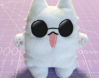 May include: A white plush toy cat wearing black sunglasses and a pink tongue sticking out. The cat has pink paw prints on its arms.