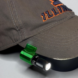 以下が含まれることがあります： オレンジとネイビーブルーで「ILLINI」の文字が刺繍されたグレーの野球帽。グリーンの懐中電灯がツバに取り付けられ、ライトが点灯しています。
