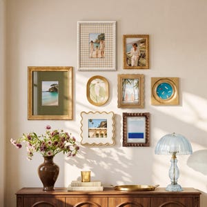 May include: A gallery wall featuring eight assorted picture frames in gold, white, and brown, a brown ceramic vase with pink flowers, a small gold candle holder, a gold tray, and a blue glass table lamp on a wooden cabinet.