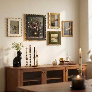 Op de afbeelding: Een houten dressoir met een zwarte vaas in de vorm van een kat met witte bloemen, vier koperen kandelaars met dinerkaarsen, een witte pompoen, een gouden pompoen en een glazen stolp. Vijf ingelijste kunstwerken hangen boven het dressoir.