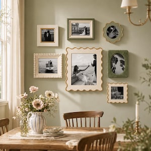 Peut inclure: Une collection de photographies encadrées de différents styles et tailles, disposées sur un mur. Les cadres sont de couleurs blanche, dorée et verte, avec des photographies en noir et blanc. Un vase de fleurs est posé sur une table en bois au premier plan.