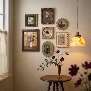 May include: A gallery wall arrangement featuring seven assorted decorative frames with floral and botanical prints, alongside a brass vase holding dark red cosmos flowers on a round wooden table, illuminated by a hanging glass pendant light.