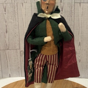 May include: A decorative figurine of a person with white hair and an eye patch, wearing a green coat with a black cape lined in red, a gold vest, and red and white striped trousers. The figure holds a small nutcracker ornament on a white string.