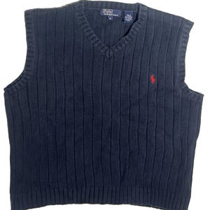 May include: A navy blue sleeveless V-neck sweater vest featuring a vertical ribbed knit pattern. The garment includes a small red embroidered logo on the left chest and a Polo by Ralph Lauren label at the inner neckline with a size M tag.