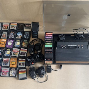 May include: A vintage Atari Video Computer System console with a clear plastic dust cover, multiple black game cartridges, four black joystick controllers with red buttons, and various connecting cables. The console features a wood-grain panel.