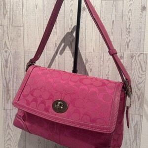 May include: A bright pink shoulder bag featuring a repeating circular logo pattern across the fabric. The bag has a fold-over flap secured with a silver-toned metal turn-lock closure, matching pink leather trim, and an adjustable shoulder strap.