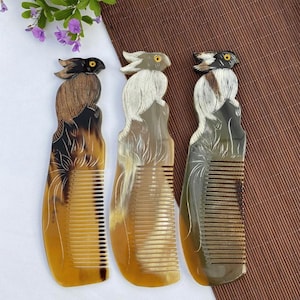 Puede incluir: Tres peines para el cabello hechos de cuerno natural, cada uno con un pájaro tallado en la parte superior del mango. Los peines muestran patrones de colores variados en tonos marrón, beige y crema, con un ojo amarillo distintivo.