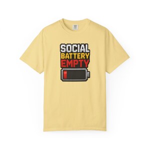 Pode incluir: Uma t-shirt de manga curta em amarelo pálido com um gráfico de pixel art no peito. O design apresenta o texto SOCIAL BATTERY EMPTY em letras de bloco brancas, amarelas e vermelhas acima de um ícone de bateria com uma barra vermelha.