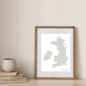 May include: A framed print featuring a gray map of Wales with place names. The print is in a wooden frame and is displayed on a light-colored surface. A stack of books and a mug are next to the frame.