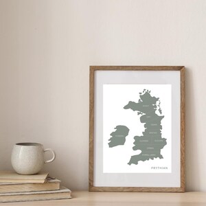 May include: A framed print featuring a map of Prythian in shades of gray. The map is labeled with place names and the word "PRYTHIAN" at the bottom. The frame is made of light brown wood. A stack of books and a ceramic mug are on the table.