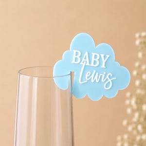 Könnte beinhalten: Ein hellblauer, wolkenförmiger Glasmarker aus Acryl mit dem weißen Schriftzug BABY Lewis. Der Marker ist so gestaltet, dass er am Rand eines klaren Glases befestigt werden kann und den Namen in einer Kombination aus Block- und Schreibschrift zeigt.