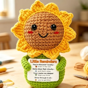 Puede incluir: Un girasol de ganchillo con una cara sonriente, pétalos amarillos y un centro marrón. Sostiene una tarjeta con el texto "Little Reminders" y citas inspiradoras. La base del girasol es verde.