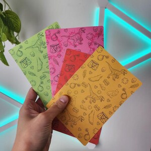 May include: A set of four rectangular cards in light green, pink, red, and yellow, each featuring a repeating black line art pattern of dinosaurs, bombs, underwear, feathers, hearts, and feet. The cards are arranged in a fan shape.