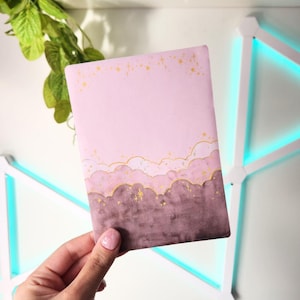 Puede incluir: Un cuaderno rosa con un diseño de acuarela de nubes en tonos morados y dorados. La parte superior del cuaderno tiene estrellas doradas. El cuaderno está sostenido en la mano de una persona.