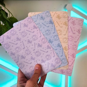 May include: Four rectangular cards featuring a repeating pattern of hockey jerseys, hockey sticks, and small hearts. The cards are coloured in shades of light purple, blue, peach, and pink, each displaying the same line-art graphic design.