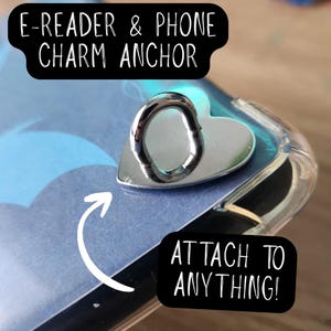 May include: A silver-colored heart-shaped metal anchor with a loop, designed for attaching charms to devices. The image includes text labels reading E-READER & PHONE CHARM ANCHOR and ATTACH TO ANYTHING, with a white arrow pointing to the anchor.