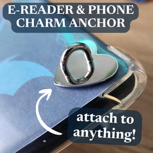 Könnte beinhalten: Ein silberner Charmanker für E-Reader und Telefone. Der Anker ist herzförmig mit einer Schlaufe zum Anbringen von Charms. Der Text "E-READER & PHONE CHARM ANCHOR" steht oben und "attach to anything!" unten.