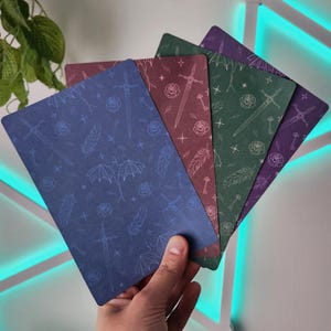 Könnte beinhalten: Ein Set aus vier rechteckigen Karten in Blau, Kastanienbraun, Grün und Violett, die jeweils ein wiederkehrendes Muster aus weißen Linienzeichnungen von Schwertern, Rosen, Federn, Schlüsseln, Sternen und Drachensilhouetten zeigen.