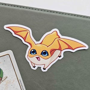 May include: A sticker featuring a cartoon character with orange wings, a yellow body, and large blue eyes. The character has a smiling expression and is set against a gray-green background. A card with Japanese text is partially visible.