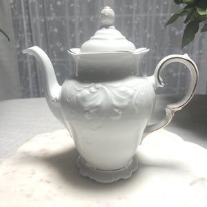 May include: A white porcelain teapot featuring an embossed scrollwork pattern on the body, a matching lid with a finial, and a curved handle and spout. The edges of the lid, handle, spout, and base are accented with a thin gold trim.