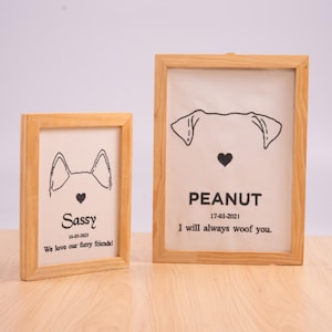 May include: Two rectangular light wood frames containing off-white fabric panels with black embroidered dog outlines, hearts, and text. The smaller frame reads Sassy 16-05-2023 We love our furry friends. The larger frame reads PEANUT 17-01-2021 I will always woof you.