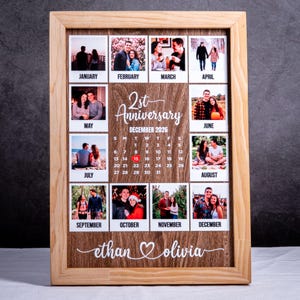 May include: A wooden framed wall calendar featuring twelve square photo prints labeled with months from January to December. The center displays the text 2nd Anniversary December 2026 with a calendar grid and the names ethan and olivia at the bottom.