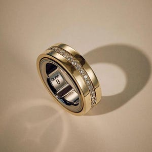 May include: A gold-toned ring with a band of small, sparkling stones. The ring has a dual-tone design, with a silver-colored interior and a gold-colored exterior. The text "OURA 8" is visible on the inner band.