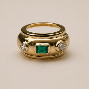 May include: A wide gold band ring featuring a central emerald-cut green gemstone flanked by two smaller clear stones, one round and one pear-shaped, set into the polished metal surface of the thick, rounded band design.