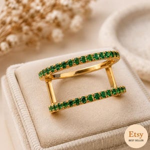 Könnte beinhalten: Ein goldener Ring mit zwei parallelen Bändern, die durch vertikale Stege verbunden sind. Jedes Band ist mit einer durchgehenden Reihe kleiner, runder, leuchtend grüner Edelsteine besetzt, was eine offene, geometrische Struktur ergibt.