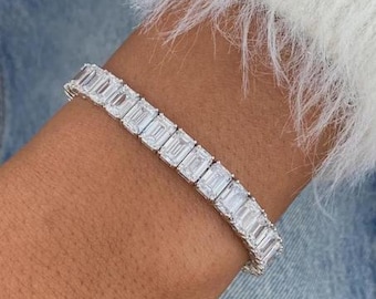 May include: A silver bracelet with rectangular, clear gemstones. The bracelet is worn on a wrist, with a white, fuzzy sleeve and blue denim in the background. The gemstones are set closely together, creating a continuous, sparkling band.