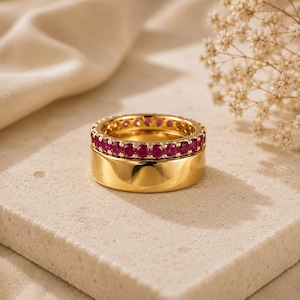 May include: A set of two stacked rings. The bottom ring is a wide, polished gold band with a smooth surface. The top ring is a thinner gold band featuring a continuous row of small, round, vibrant pink gemstones set in a prong setting.
