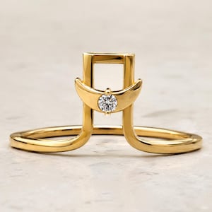 May include: A gold ring featuring a vertical rectangular frame structure with a crescent moon motif centered at the base of the frame. A single round, clear, faceted gemstone is set in the middle of the crescent moon, creating a distinct architectural design.
