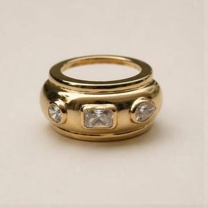 Peut inclure: Une bague dorée ornée de trois pierres précieuses transparentes. La bague a une bande épaisse et une forme arrondie, avec une pierre rectangulaire au centre et deux pierres plus petites, de formes différentes, de chaque côté. La bague est probablement en métal doré.