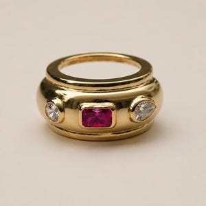 Op de afbeelding: Een brede gouden ring met een centrale rechthoekig geslepen felroze edelsteen, geflankeerd door twee heldere, ronde en peervormige stenen. Het gepolijste gouden oppervlak heeft een glad, afgerond profiel met een hoogglans afwerking.