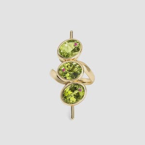 Puede incluir: Un anillo de oro con tres piedras preciosas ovaladas de color verde claro, cada una acentuada con una pequeña piedra roja. El anillo tiene un diseño único con las piedras dispuestas verticalmente y conectadas por una banda curva.