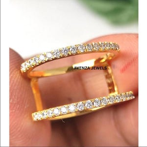 May include: A double-band ring with a gold band and a row of small, clear gemstones. The ring has the text "AVENZA JEWELS" on the side. The ring is held by a person's hand.