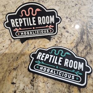 May include: Two black rectangular signs with rounded edges featuring the text REPTILE ROOM and HOGALICIOUS. One sign has peach-coloured snake and leaf graphics, while the other has teal-coloured snake and leaf graphics. Both signs have white borders.