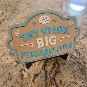 Op de afbeelding: Een beige, rechthoekig bord met afgeronde randen en een lichtblauwe omlijning. Het bord bevat een wit hersenpictogram, twee kleine sterren en de tekst "TINY BRAINS, BIG PERSONALITIES" in verhoogde lichtblauwe en witte letters op een zwarte standaard.