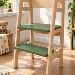 May include: A light-colored wooden learning tower featuring two horizontal steps painted in a matte olive green finish. The structure has a tall back support, open side panels with rectangular cutouts, and four sturdy legs for stability.