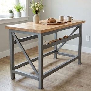 Può includere: Un'isola da cucina rettangolare con un spesso piano di lavoro in legno chiaro e una robusta struttura in metallo grigio con supporti laterali a forma di X. La base include un ripiano orizzontale che ospita un martello e un tagliere.