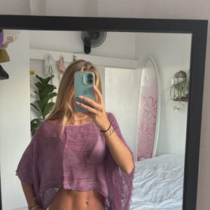 Puede incluir: Un top corto de color morado con mangas cortas, confeccionado en una tela texturizada semitransparente con bordes deshilachados, combinado con pantalones vaqueros blancos. El top presenta un cuello redondo ancho y un corte holgado.