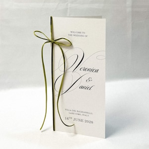 May include: A tall, folded white wedding programme card featuring black text that reads "WELCOME TO THE WEDDING OF Veronica & Daniel VILLA DEL BALBIANELLO, LAKE COMO, ITALY 16TH JUNE 2028". A thin, olive green velvet ribbon is tied in a bow on the left side.