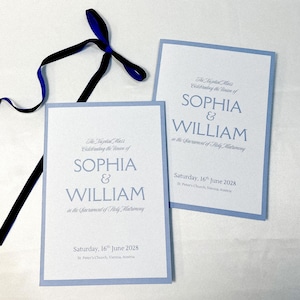 May include: Two rectangular wedding programmes with light blue borders and white cardstock. The text reads: The Nuptial Mass Celebrating the Union of SOPHIA & WILLIAM in the Sacrament of Holy Matrimony, Saturday, 16th June 2028, St. Peter's Church, Vienna, Austria. A dark blue ribbon is tied above.