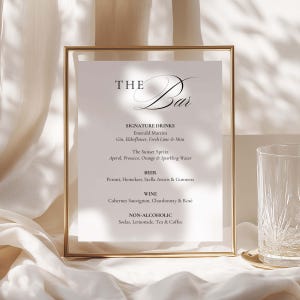 Może przedstawiać: Prostokątna złota metalowa ramka zawierająca białą kartę menu z napisem THE Bar. Menu zawiera autorskie drinki, takie jak Emerald Martini i The Sunset Spritz, a także sekcje z piwem, winem i napojami bezalkoholowymi zapisane czarną czcionką.