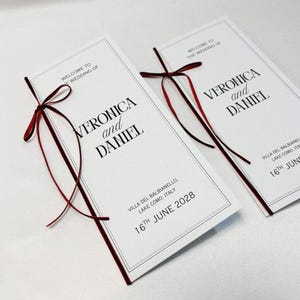 Può includere: Due programmi di matrimonio rettangolari bianchi con testo nero che recita "WELCOME TO THE WEDDING OF VERONICA and DANIEL" e "VILLA DEL BALBIANELLO, LAKE COMO, ITALY 16TH JUNE 2028". Ogni programma presenta un sottile nastro rosso scuro.