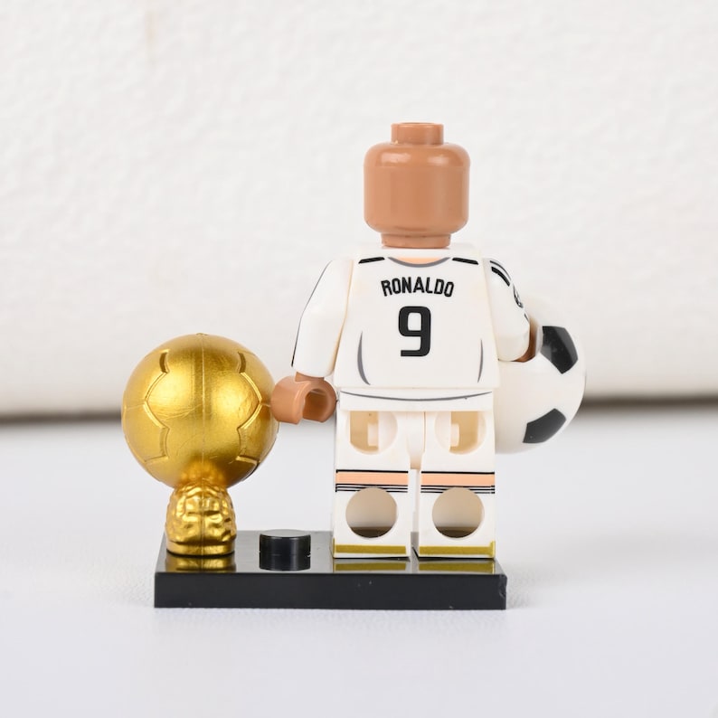 May include: A Lego figure of a footballer in a white kit, with "Ronaldo" and the number "9" on the back. The figure holds a football and stands beside a gold trophy.