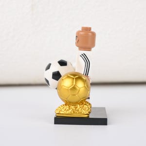 May include: A Lego footballer figurine in a white kit with black stripes, holding a black and white football and a gold trophy. The figurine is on a black base.