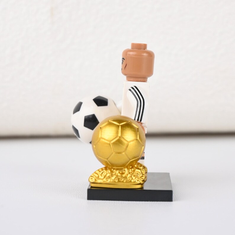 May include: A Lego footballer figurine in a white kit with black stripes, holding a black and white football and a gold trophy. The figurine is on a black base.