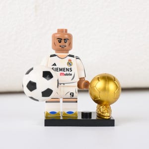 May include: A Lego football player figurine in a white kit with black detailing, holding a football and a golden trophy. The kit features the number 9 and the Siemens Mobile logo. The figurine is on a black base.