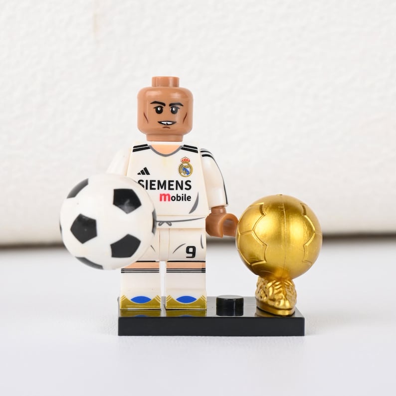 May include: A Lego football player figurine in a white kit with black detailing, holding a football and a golden trophy. The kit features the number 9 and the Siemens Mobile logo. The figurine is on a black base.